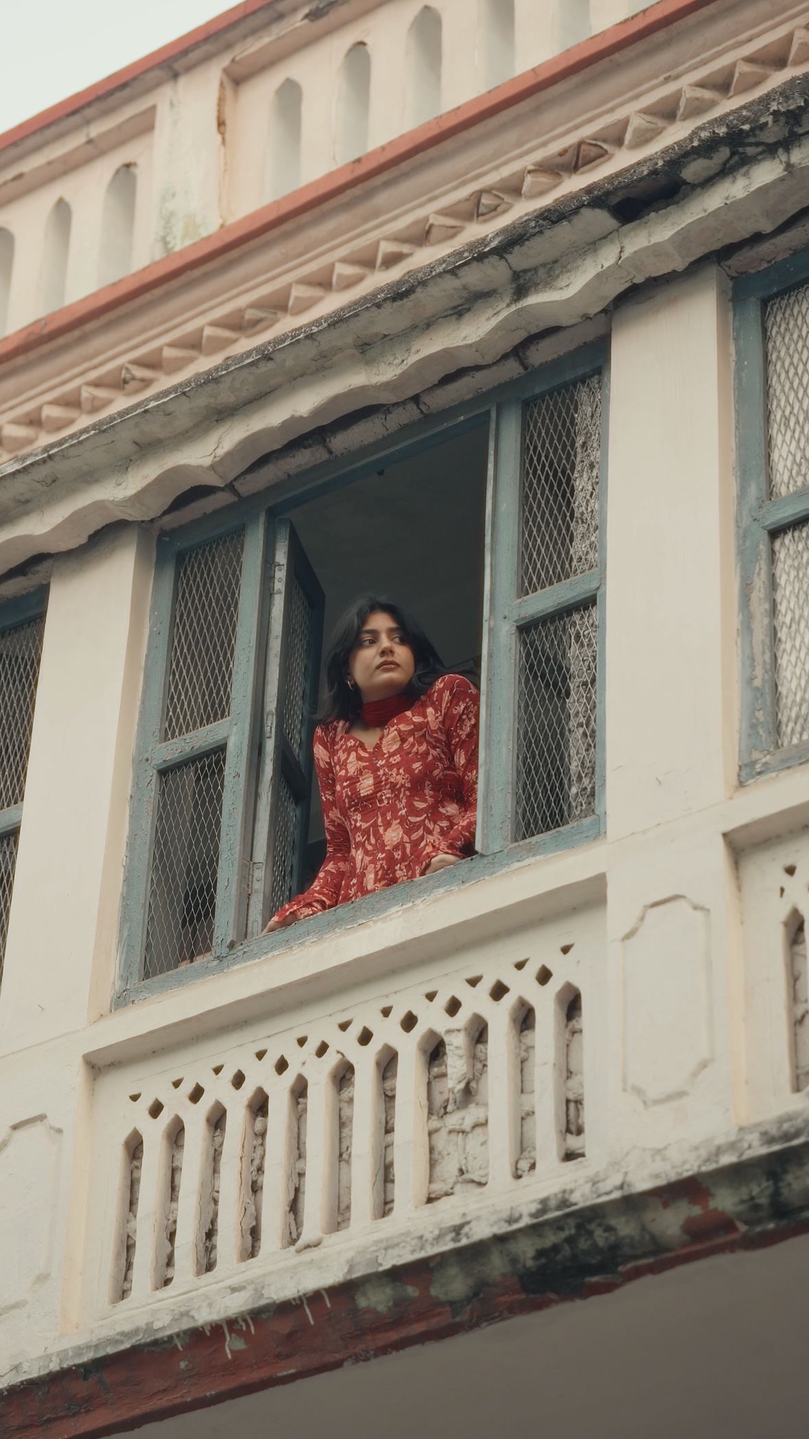 A woman in a red floral kurta leaning out of an old balcony window
