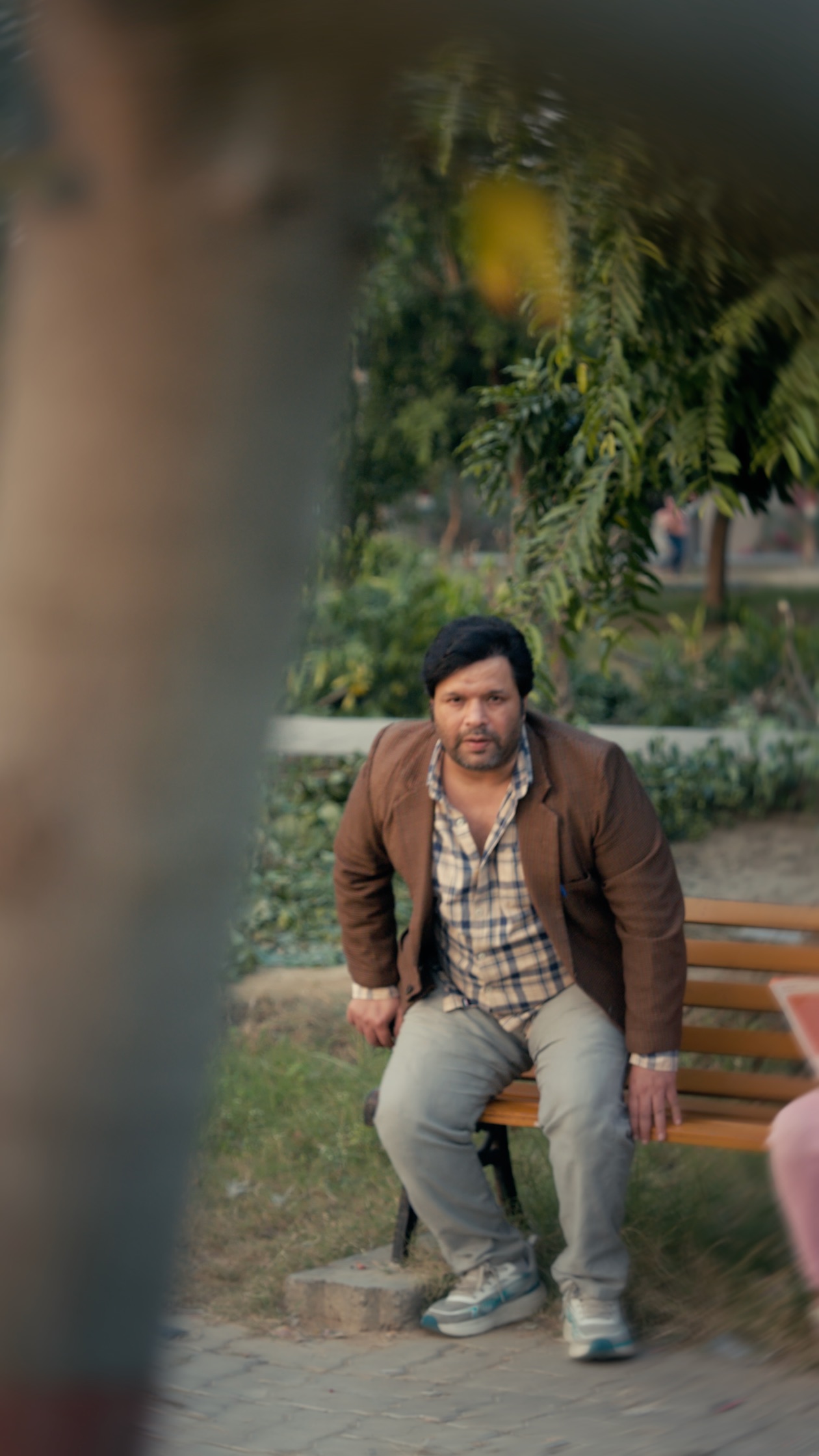 A man seated on a park bench, shot through an out-of-focus foreground