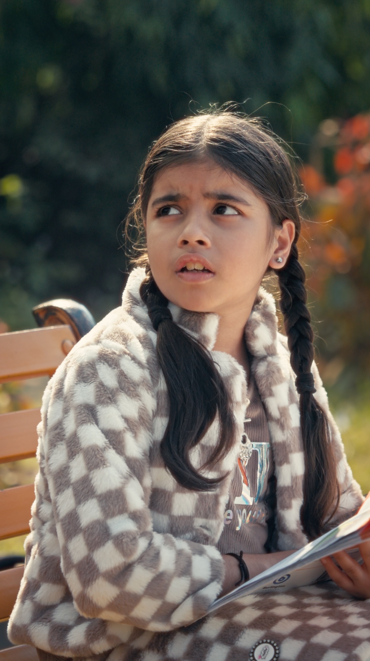 A young girl in a checkered jacket, caught mid-reaction on a park bench
